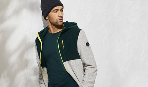 Ein Mann steht mit einer Fleecejacke in grau an einer Mauer.