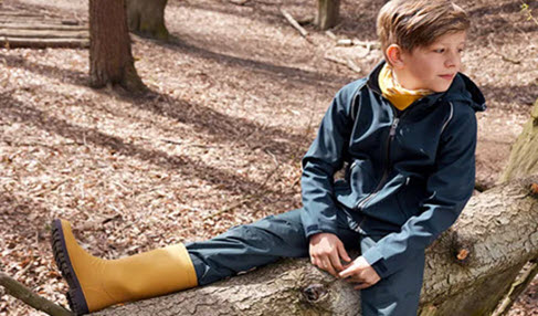 Sicher vor Matsch: Ein Junge sitzt in seiner Regenhose und gelben Gummistiefeln auf einem Baumstamm.