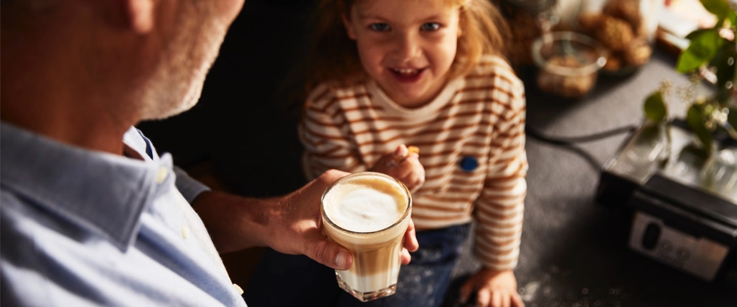 Ein kleines Mädchen schaut freudig und neugierig zu einem Mann hoch, der einen Kaffee trinkt.