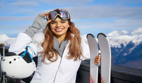 Eine Frau lächelt und winkt in die Kamera. Sie trägt eine weiße Ski Jacke, graue Handwärmer und einen grauen Schlauchschal.