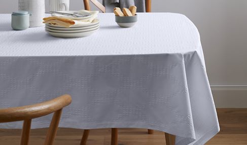 Ein Esstisch, auf dem eine Tischdecke aus Leinen liegt. Tisch und Wohnbereich sind in einem natürlich-rustikalen Stil gehalten, zu dem dieser Stoff bestens passt.
