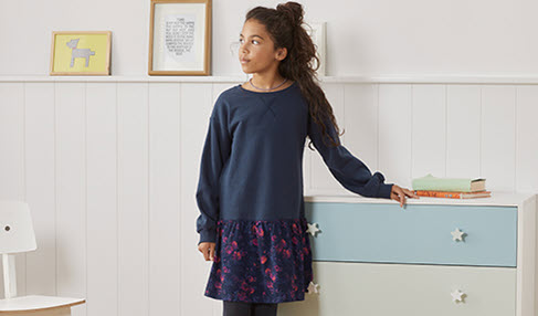 Immer warm bleiben: Ein Mädchen steht mit langem Shirt in blau und Rock in die Kinderzimmer.