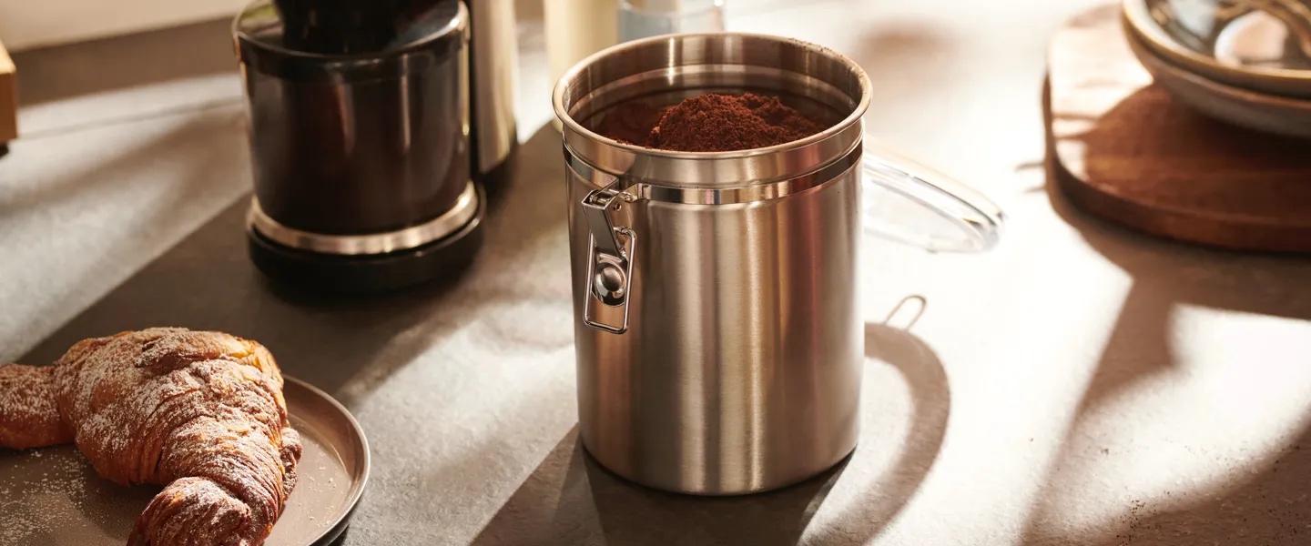 Kaffeebohnen werden in eine Kaffeedose verkippt. Dieses stimmungsvolle Kaffeegenussbild leitet den Schwerpunkt der Kaffeeaufbewahrung ein.