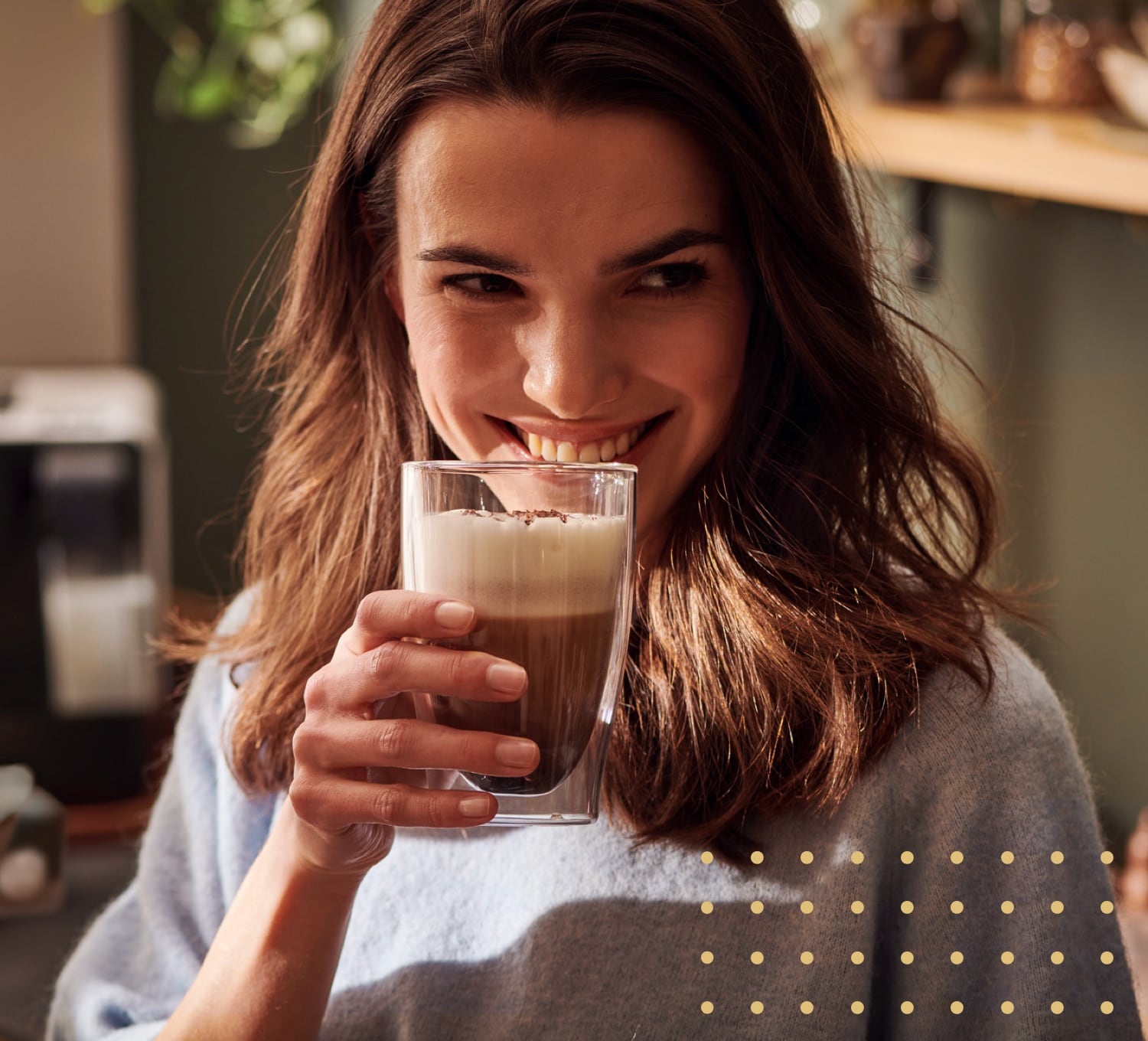 Eine Frau trinkt ein Kaffee-Milchgetränk aus einem Latte Macchiato Glas und lächelt dabei