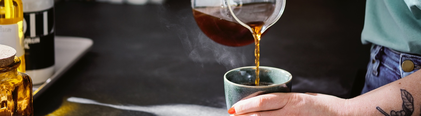 Eine Person steht an einer Küchentheke und schenkt sich Specialty Coffee Filterkaffee aus einer Glaskanne in einen Kaffeebecher ein.