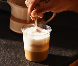 Jemand rührt in einem Glas einen Kaffee mit Milchschaum um