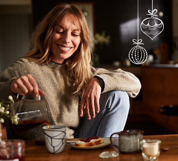 Eine Frau sitzt entspannt an einem Tisch und schenkt sich Kaffee in eine Tasse ein.