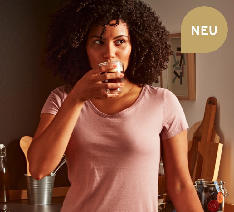 Eine Frau steht in einer Küche und trinkt einen Caffè Crema aus einem Kaffeeglas.