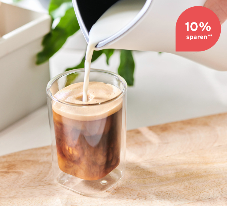 Milchschaum wird aus einem Milchaufschäumer in ein Caffè Crema Glas gegossen.