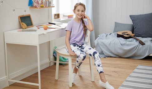 Ein Mädchen sitzt an dem Schreibtisch in ihrem Kinderzimmer. Im Hintergrund steht ihr Kinderbett.