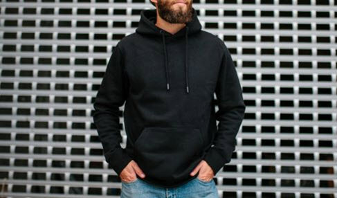 Vor einer gefliesten Wand steht ein Mann in schwarzem Kapuzenpullover mit Händen in der Bauchtasche.