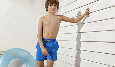Stylische Farben. Ein Junge trägt eine blaue Badehose und lehnt sich lässig gegen eine Hauswand.