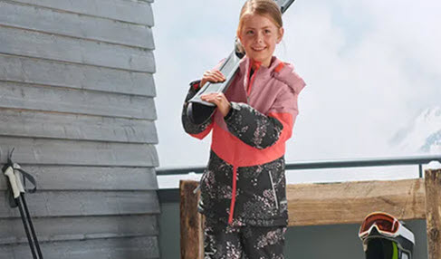 Ein Mädchen trägt eine rosa-rote Regen- und Schneejacke und hält Skier in der Hand.